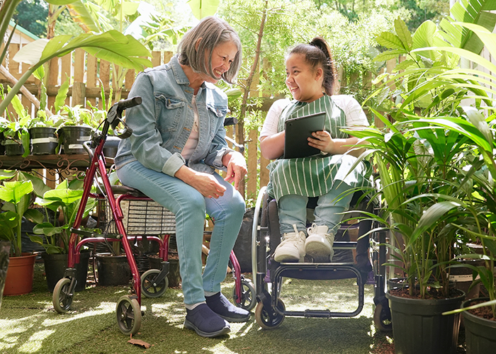 Two smiling women with various disabilities looking at digital tablet and talking at plant nursery